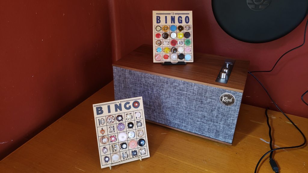 Photo of corner of Trixie's brick-colored cabin and vintage wooden desk featuring bingo cards with antique glass and rhinestone buttons and mid-century modern Klipsch speaker