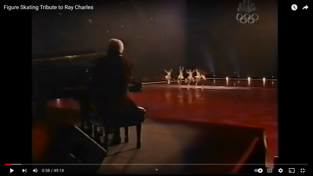 Still image from TV special Figure Skating Tribute to Ray Charles shows Ray from behind playing grand piano on elevated stage with expanse of ice performance area with group of ice dancers down-field with short skirts floating and arms raised jubilantly